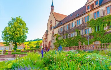 Blick aus dem blühenden Klostergarten auf das Kloster Bronnbach. Die Fassade ist begrünt. Im Hintergrund sind Steinfiguren und ein großer Baum zu sehen. | © Liebliches Taubertal