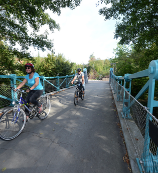 Radfahrer fahren über eine Brücke auf der Wanderbahn von Mudau nach Mosbach. Die Brücke hat ein blaues Geländer und ist von Bäumen umgeben. | © Touristikgemeinschaft Odenwald e.V.