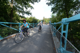 Radfahrer fahren über eine Brücke auf der Wanderbahn von Mudau nach Mosbach. Die Brücke hat ein blaues Geländer und ist von Bäumen umgeben. | © Touristikgemeinschaft Odenwald e.V.