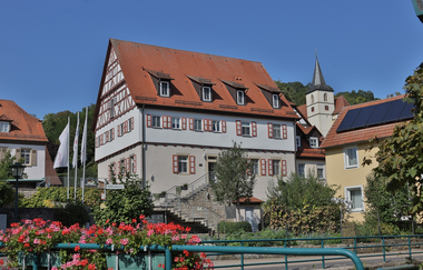 Ein altes Fachwerkgebäude und eine Kirche sind von einer Brücke aus zu sehen | © Touristikgemeinschaft Hohenlohe e. V. | Andi Schmid