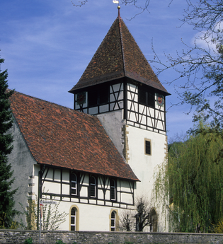 Die Kirche hat Fachwerk rund ums Gebäude und ist von Bäumen umgeben | © Touristikgemeinschaft Hohenlohe e. V. | Eva Maria Kraiss
