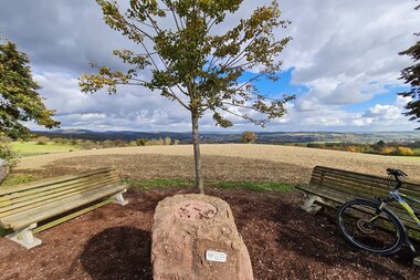 Fernblick zum  Odenwald und über das Kraichgauer Hügelland. Im Vordergrund ein symbolischer Grenzstein "Odenwald - Kraichgau", zwei Sitzbänke und ein Fahrrad. | © Landratsamt Rhein - Neckar - Kreis