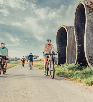 Radler fahren auf einem Fahrradweg, daneben befindet sich eine Skulptur, die eine Straße in Wellen zeigt; im Hintergrund befinden sich Wiesen und Wälder | © Touristikgemeinschaft Odenwald e.V.