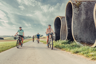 Radler fahren auf einem Fahrradweg, daneben befindet sich eine Skulptur, die eine Straße in Wellen zeigt; im Hintergrund befinden sich Wiesen und Wälder | © Touristikgemeinschaft Odenwald e.V.