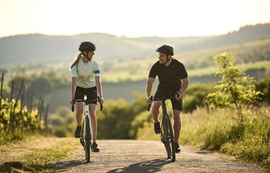 Zwei Radfahrernde fahren mit Helmen auf einer kleinen Straße durch die Weinberge im Kraichgau-Stromberg. Im Hintergrund liegen sanfte Hügel im warmen Abendlicht. | © Tourimia Tourismus GmbH | Stefan Leitner Photography