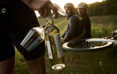 Radpause in den Weinbergen: Eine Person hält eine Weinflasche und zwei Gläser, im Hintergrund sitzen zwei Radfahrer*innen mit Helmen neben ihren Rädern im Grünen. | © Tourimia Tourismus GmbH | Stefan Leitner Photography