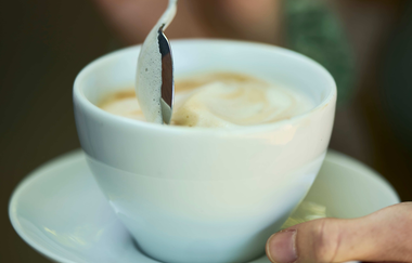 Hand hält eine Tasse Cappuccino, während der Milchschaum mit einem Löffel umgerührt wird. | © Tourimia Tourismus GmbH | Stefan Leitner Photography