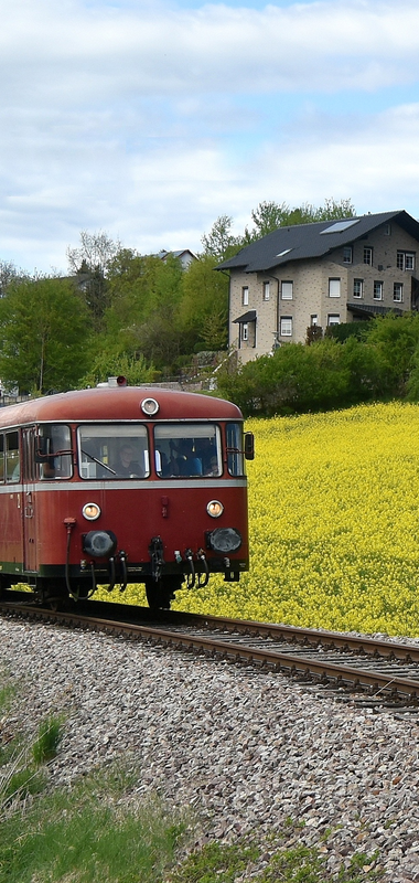 Zug mit zwei Waggons fährt auf Gleisen neben einem blühenden Feld und mehreren Häusern im Hintergrund | © Willi Hofmann