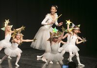 Eine Gruppe von Kindern in weißen Ballettkostümen mit Sternen in der Hand und eine ältere Tänzerin mit Stab auf einer Bühne vor schwarzem Hintergrund