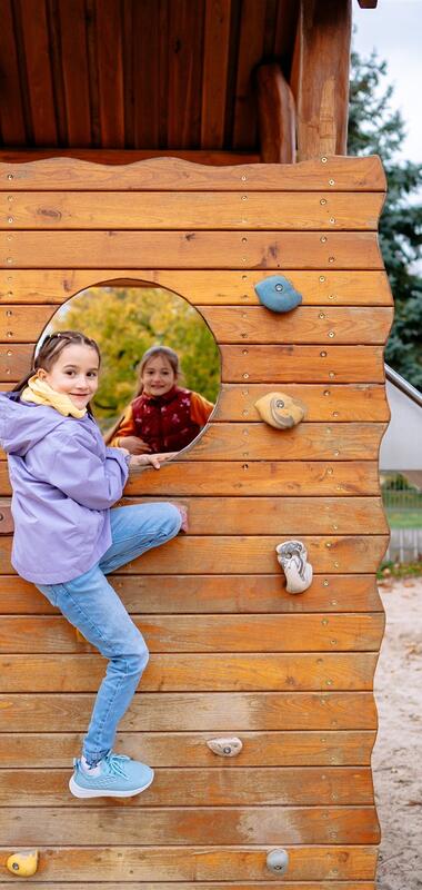 Kind klettert an einer Holz-Kletterwand mit bunten Griffen auf einem Spielplatz, ein weiteres Kind schaut durch ein rundes Fenster in der Wand | © Landratsamt Rhein-Neckar-Kreis; Foto Sebastian Weindel
