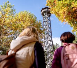Zwei Personen mit Rucksäcken blicken auf einen hohen Aussichtsturm aus Metall, umgeben von herbstlich gefärbten Bäumen. | © Landratsamt Rhein-Neckar-Kreis, Sebastian Weindel